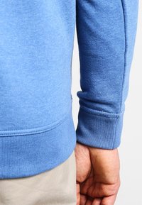 Blå sweatshirtärm med ribbad mudd; blandmaterial av bomull. Slät textur, avslappnad design med minimalistisk stil och inga synliga mönster.