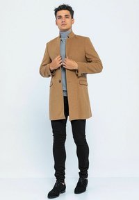 Beige Wollmischung Mantel mit Stehkragen, zwei vorderen Taschen und tonalen Knopfdetails, kombiniert mit einem hellgrauen Rollkragenpullover und schwarzen Hosen.