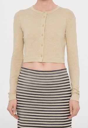 Personne portant un top beige à manches longues raccourci avec boutons et une jupe tricotée à rayures taille haute dans des tons neutres, debout sur un fond blanc.