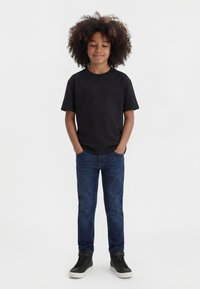 Otrok s kodrastimi lasmi, oblečen v črno majico, modre jeans hlače in črne superge, stoji z rokami v žepih na enostavni belem ozadju.