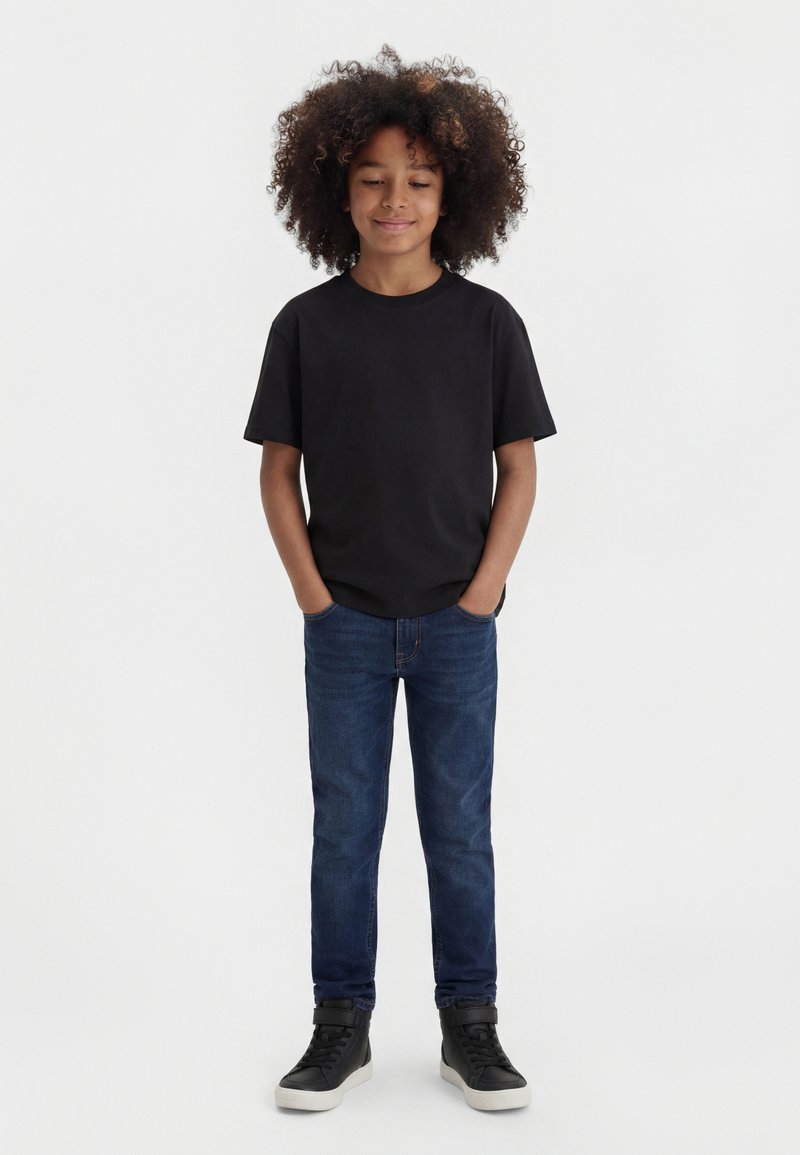 Otrok s kodrastimi lasmi, oblečen v črno majico, modre jeans hlače in črne superge, stoji z rokami v žepih na enostavni belem ozadju.