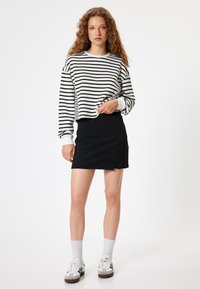 Schwarzer Rock kombiniert mit einem gestreiften Langarmshirt mit weißen Manschetten. Das Model trägt weiße Socken und Sneakers. Minimalistisches Design.