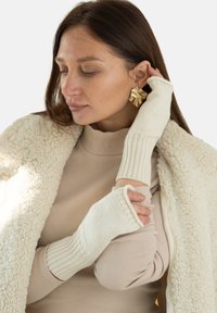 Kremowe, dziergane rękawiczki bez palców z prążkowanymi mankietami, noszone przez osobę w beżowej, prążkowanej bluzce i puszystym, białym płaszczu.