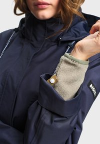 Personne ajustant le poignet de sa veste bleu marine sur une manche en polaire beige, montrant les cordons de serrage de la veste et le patch avec le logo "AIGLE" sur la poitrine.