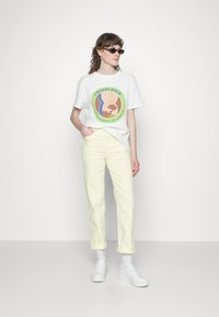 T-shirt gráfica branca com um design colorido, combinada com calças amarelas claras com as bainhas dobradas e ténis brancos. Óculos de sol completam o visual.
