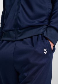 Giacca e pantaloni da track di colore navy realizzati in un tessuto morbido e elastico; presentano il logo a chevron bianco sui pantaloni, una zip frontale e polsini aderenti.