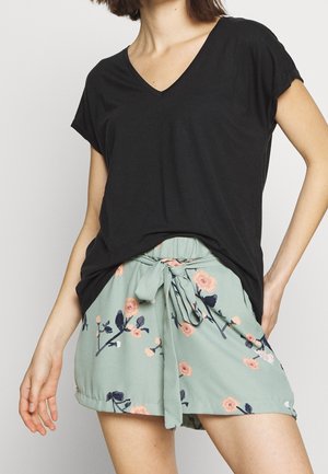 Frau trägt ein schwarzes V-Ausschnitt-Kurzarmtop und hellgrüne, geblümte Shorts mit vorderem Bindegürtel, steht mit einer Hand in der Hüfte.