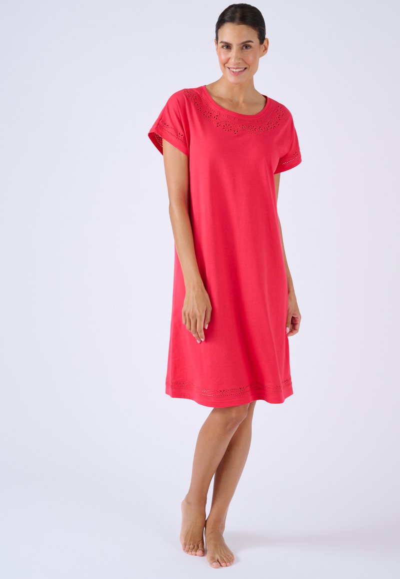 Mujer descalza de pie, vistiendo un vestido rosa brillante hasta la rodilla con detalles florales recortados en el escote y las mangas.