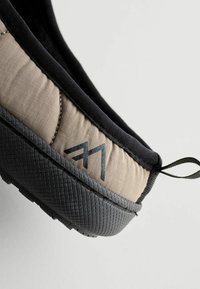 Zapato tipo slip-on con un upper de textil beige, ribetes negros y suela de goma negra texturizada. Presenta un logotipo en el lateral y una tira para facilitar el calzado.