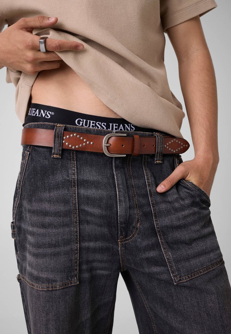 Personne portant un jean en denim foncé avec une ceinture cloutée marron et une ceinture noire portant l’étiquette "GUESS JEANS", visible sous une chemise beige.