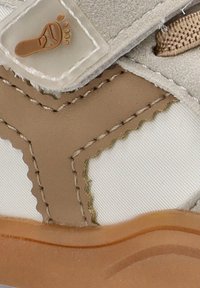 Zapato infantil beige con tejido texturizado, panel blanco contrastante y suela marrón. Presenta un logo de pie y acentos cosidos.