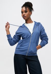 Μπλε φούτερ με φερμουάρ, κατασκευασμένο από μαλακό ύφασμα, με κουκούλα, μπροστινές τσέπες και ριμπ μανσέτες. Συνδυάζεται με λευκή μπλούζα και σκούρο τζιν.