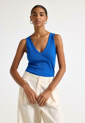 Kvinde iført en ærmeløs blå V-hals top og cremefarvede bukser med høj talje, stående med hænderne foldet foran mod en ensfarvet baggrund.