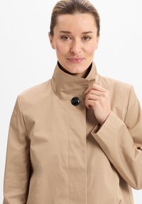 Beige, met kraag, jas gemaakt van een glad materiaal. Heeft een opvallende zwarte knop en een minimalistisch ontwerp met strakke lijnen.