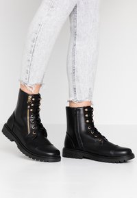 Svarta ankelstövlar i konstläder med rund tå, guldfärgade öljetter, dragkedjedetalj och texturerad gummisula. Bärs med slitna grå jeans.