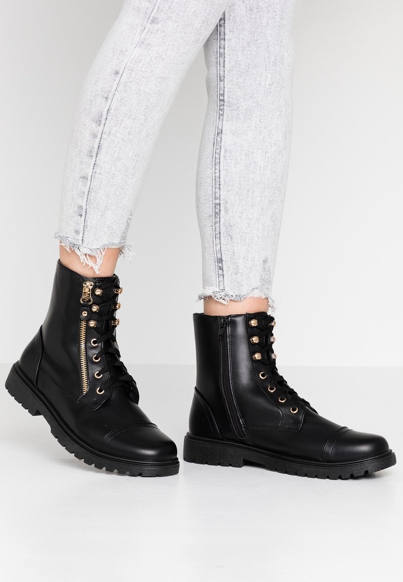 Svarta ankelstövlar i konstläder med rund tå, guldfärgade öljetter, dragkedjedetalj och texturerad gummisula. Bärs med slitna grå jeans.