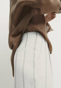 Blouse marron transparente portée sur un pantalon blanc à rayures fines verticales. Le pantalon possède une poche latérale et une coupe ajustée.