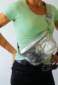 Zilveren metallic heuptas met een gebogen ontwerp, ritssluiting en verstelbare gestreepte schouderband. Draagband rond de taille over een groene shirt.