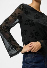 Top nero trasparente con motivo floreale in velluto, maniche lunghe a campana, scollo rotondo e materiale liscio. Indossato sopra dei jeans, con le mani posate sui fianchi.