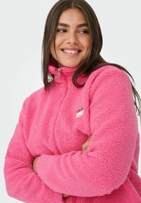Kurtka polarowa w jaskrawym różowym kolorze, z wysokim kołnierzem, zamkiem błyskawicznym i teksturowanym, przytulnym materiałem. Zawiera małą naszywkę z logo.