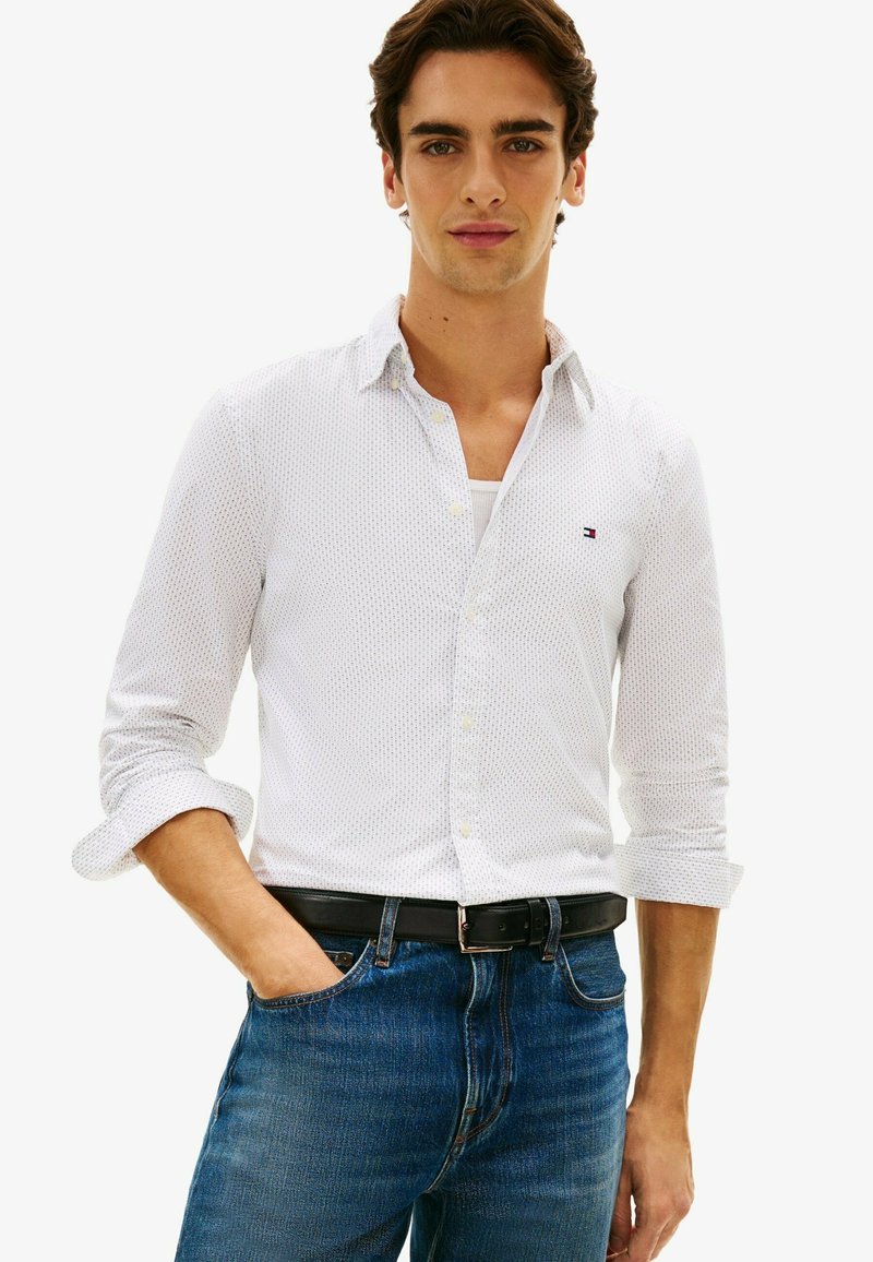 Jeune homme aux cheveux foncés portant une chemise blanche à motifs boutonnée, manches retroussées, jean bleu et ceinture noire, main dans la poche.