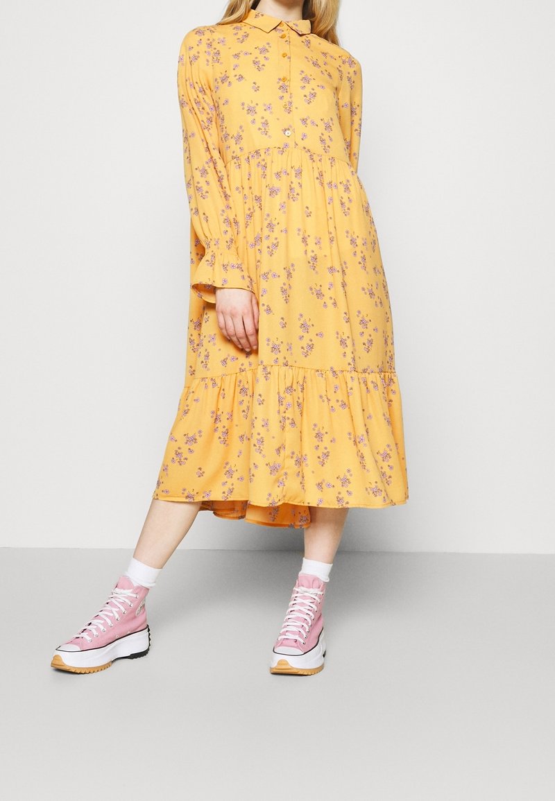 Femme portant une robe midi jaune à fleurs avec des manches longues et des baskets montantes roses avec semelles et lacets blancs.