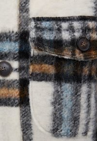 Wollhemd mit einem karierten Muster in Schwarz, Blau und Orange, einer grauen Tasche und einem dunklen Knopf. Strukturierter Stoff mit einer pelzigen Optik.