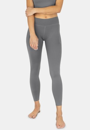 Frau trägt graue Leggings mit hoher Taille und passendem Croptop, steht barfuß mit einem Arm über dem Oberkörper verschränkt.