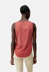 Rode mouwloze top met een gladde textuur, afgeronde zoom en een semi-doorzichtige achterkant. Gecombineerd met beige leggings met subtiele stiksels.