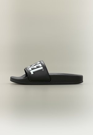 Sandale slide noire avec une large bride et le logo blanc "DIESEL" sur la bride, vue de profil sur un fond beige uni.