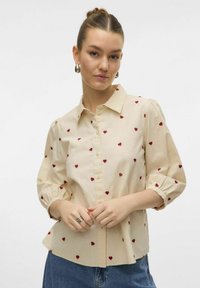 Beige knapp-up skjorta med korta puffärmar, prydd med små röda hjärtan, och en böjd fåll. Texturerad bomullstyg.