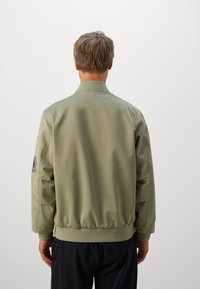 Man facing away wearing a light olive green bomber jacket with ribbed cuffs and waistband, paired with black pants.