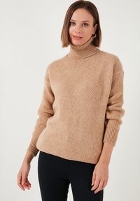 Bež turtleneck pulover od mekog pletenog materijala, sa rebrastim manžetama i opuštenim krojem. Nosi se uz tamne traperice.