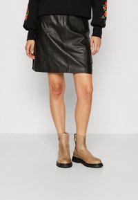 Botas de cuero beige hasta el tobillo con suelas de goma negra, cremallera lateral y un acabado suave, combinadas con una falda mini de cuero negra.