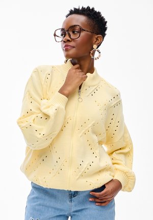 Kobieta o krótkich kręconych włosach, nosząca duże okulary w oprawkach w kształcie żółwia, kolczyki koła, żółty sweter rozpinany na zamek wykonany na drutach oraz niebieskie dżinsy, pozująca zamyślona.