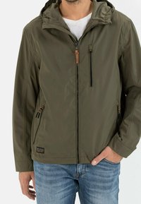 Veste légère vert olive avec capuche, dotée d'une fermeture éclair à l'avant, de deux poches latérales et d'un accent en cuir marron sur la tirette de la fermeture.