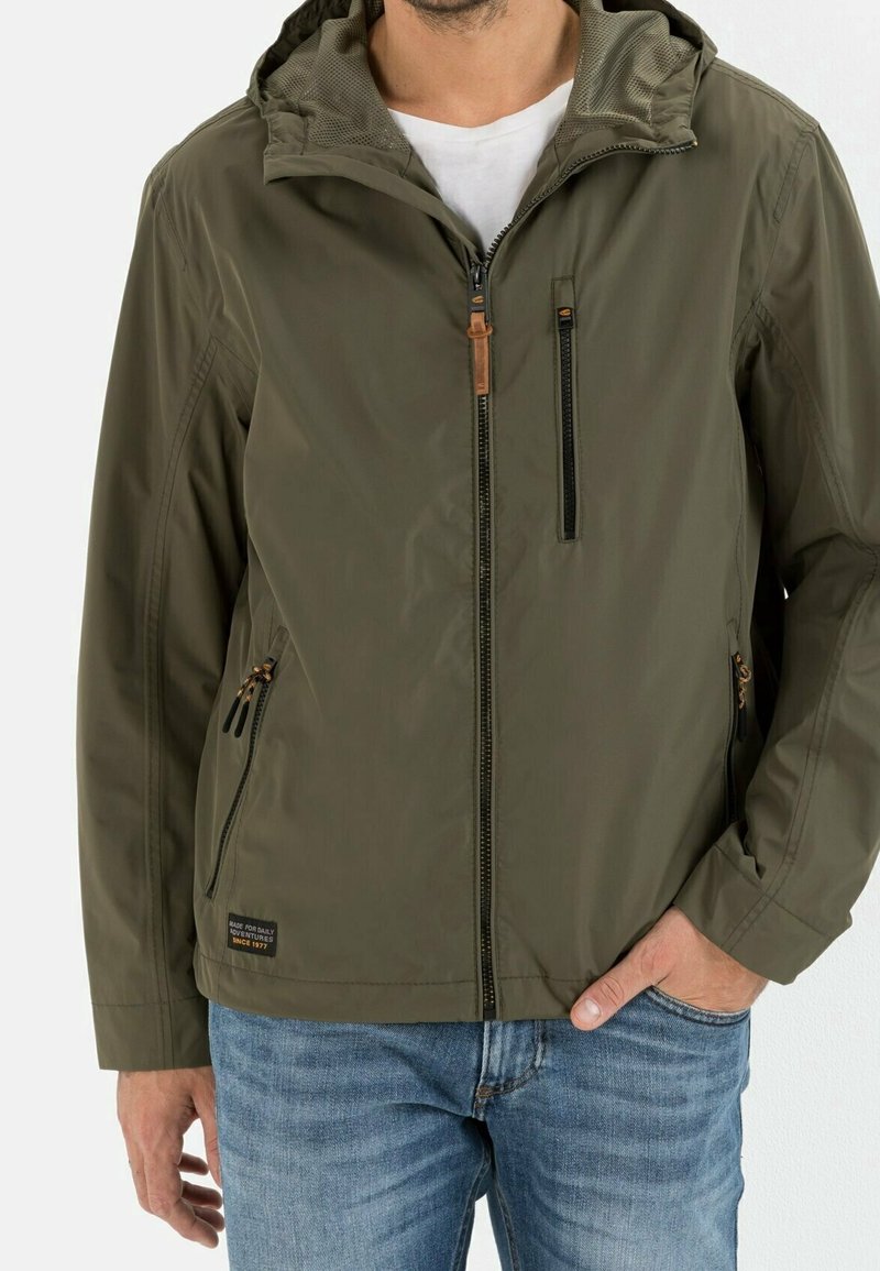 Veste légère vert olive avec capuche, dotée d'une fermeture éclair à l'avant, de deux poches latérales et d'un accent en cuir marron sur la tirette de la fermeture.