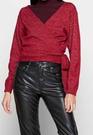 Femme portant un pull cache-cœur rouge noué sur le côté, par-dessus un col roulé sombre et un pantalon en cuir noir, se tenant devant un fond uni.