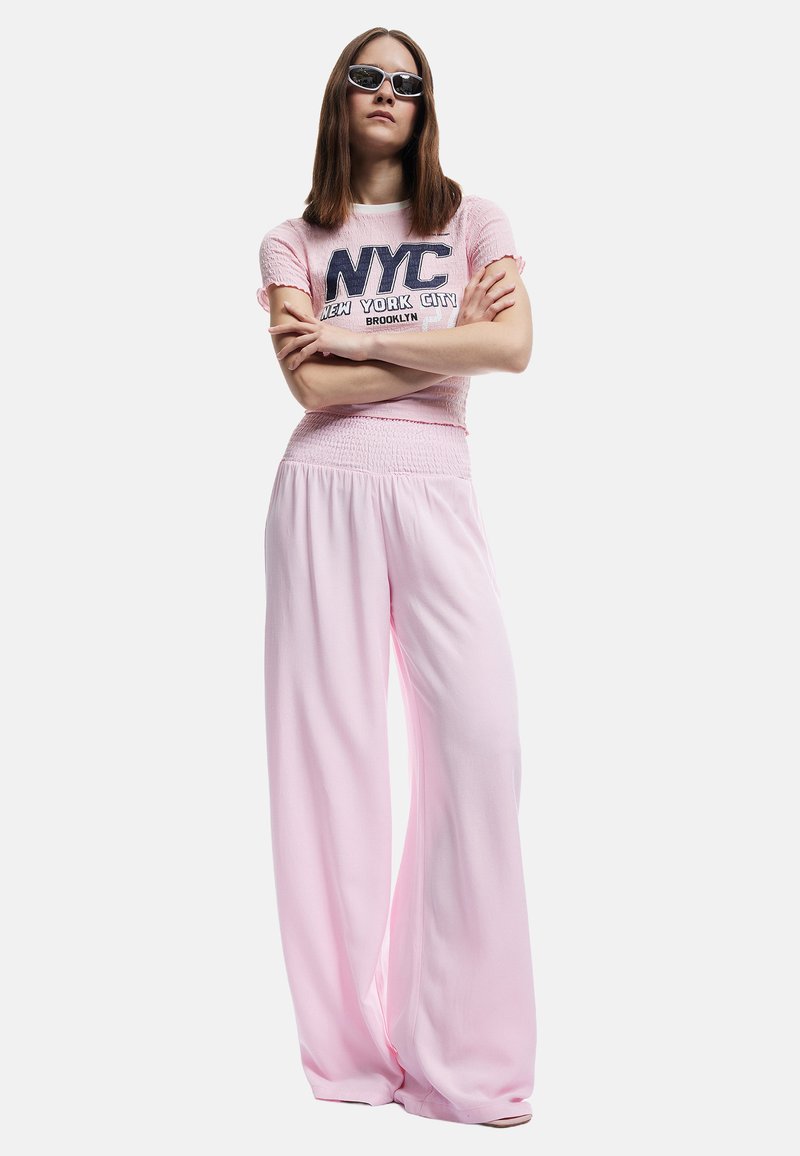 Rozewijd gelakte broek met een smocktail waistband, gecombineerd met een lichtroze NYC graphic t-shirt. Model draagt zwarte zonnebril.
