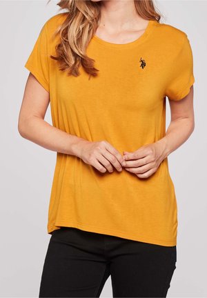 Femme portant un T-shirt jaune moutarde à manches courtes avec un petit logo noir brodé sur la poitrine gauche, associé à un pantalon noir.