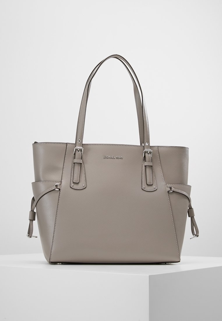 Sac cabas taupe Michael Kors avec double poignées, sangles latérales à boucle et logo de la marque visible sur une surface en cuir lisse.