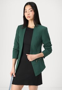 Zelený sakou s elegantním designem, přiléhavým střihem, dlouhými rukávy a zipovými detaily v pase. Spárováno s černými šaty.