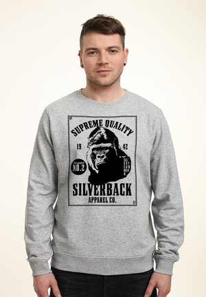 Młody mężczyzna w szarej bluzie z czarną grafiką goryla i napisem „Supreme Quality Silverback Apparel Co.”