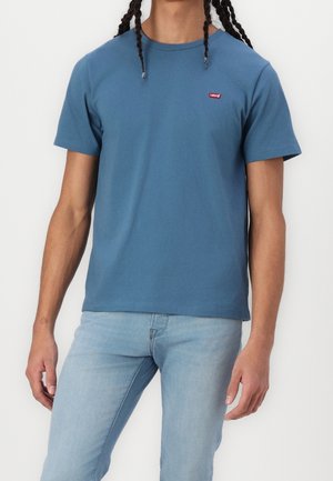 Homme portant un t-shirt Levi's bleu et un jean bleu clair, debout devant un fond blanc, cadré au-dessus du cou.