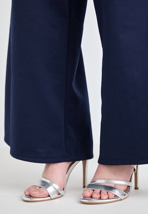 Pieds dans des sandales argentées à talons hauts avec des brides croisées, portées avec un pantalon large bleu foncé.