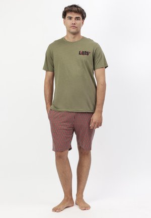 Hombre de pie descalzo vistiendo una camiseta de manga corta verde oliva con el logo "LOIS" y pantalones cortos a rayas rojas y negras.