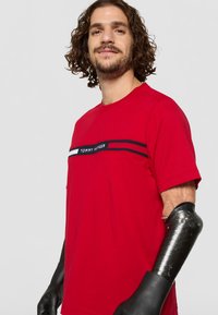 Kortsiooniga mees lokkis juustega, seljas punane Tommy Hilfiger T-särk ja mustad proteeskäed kinnastega, seismas ühtlase tausta ees.