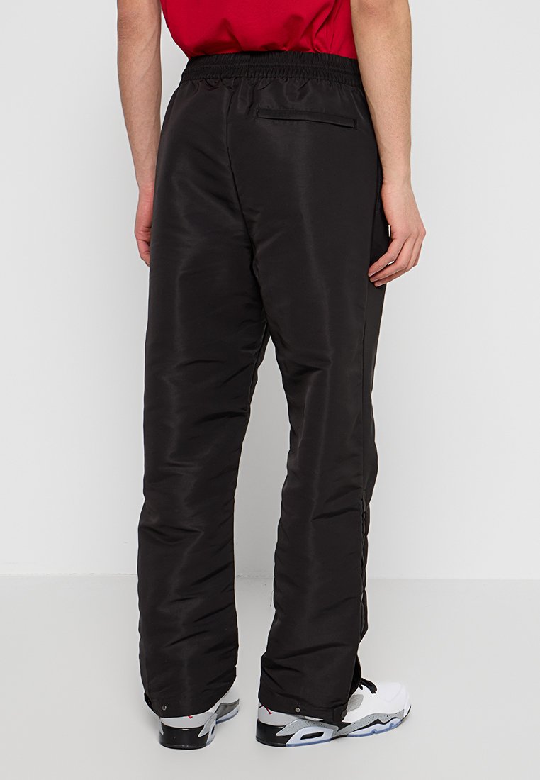 Schwarze gepolsterte Hosen mit einem elastischen Bund, ausgestattet mit einer Gesäßtasche und einer glatten, glänzenden Textur. Getragen mit weißen Turnschuhen.