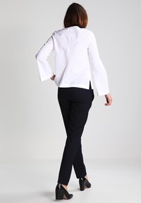 Blusa di cotone bianca con maniche a campana, orlo a strati, abbinata a pantaloni neri aderenti e scarpe nere con tacco alto, vista da dietro.