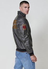 Chupa de bomber gris con cuello alto, que presenta texto bordado y parches. Lleva puesta sobre jeans de mezclilla azul. Textura suave con puños acanalados.